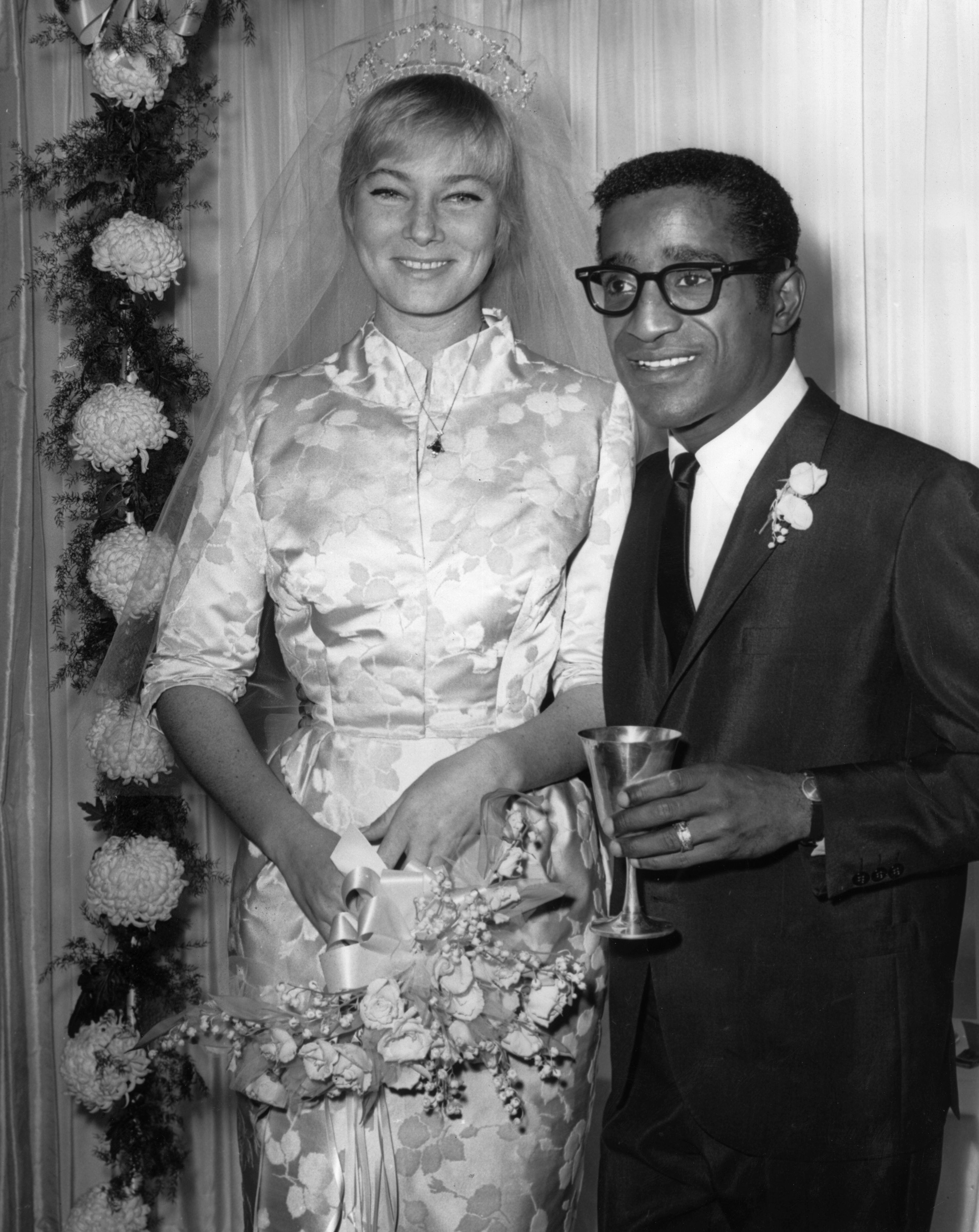 13th November 1960: American actor, singer and dancer Sammy Davis Jr. (1925 - 1990) stands next to his wife, Swedish actor May Britt, at their wedding reception at Davis's home, Hollywood, California. Britt wears a silk brocade wedding gown with a veil and bouquet. Davis wears a suit and holds a goblet. (Photo by Hulton Archive/Getty Images)