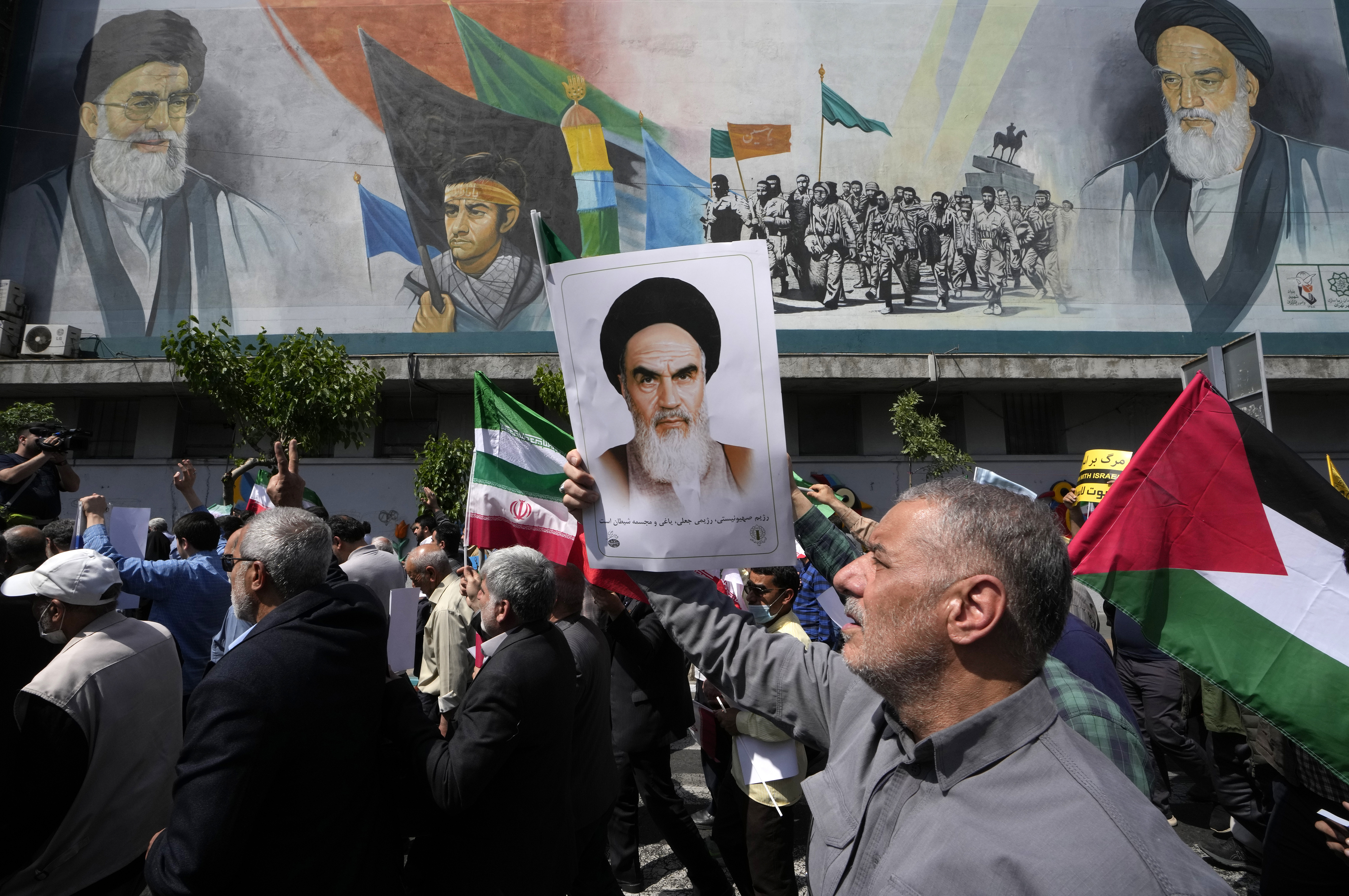 Iranian worshippers walk past a mural showing the late revolutionary founder Ayatollah Khomeini, right, Supreme Leader Ayatollah Ali Khamenei, left, and Basij paramilitary force, as they hold a poster of Ayatollah Khomeini and Iranian and Palestinian flags in an anti-Israeli gathering after their Friday prayer in Tehran, Iran, Friday, April 19, 2024.