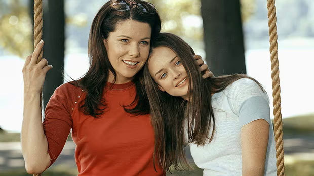 The main characters from the Gilmore Girls, mother Lorelai Gilmore and her daughter Rory, sit outside on a swing together. Lorelai is wearing a red t-shirt and Rory is a light blue top. They are smiling. 