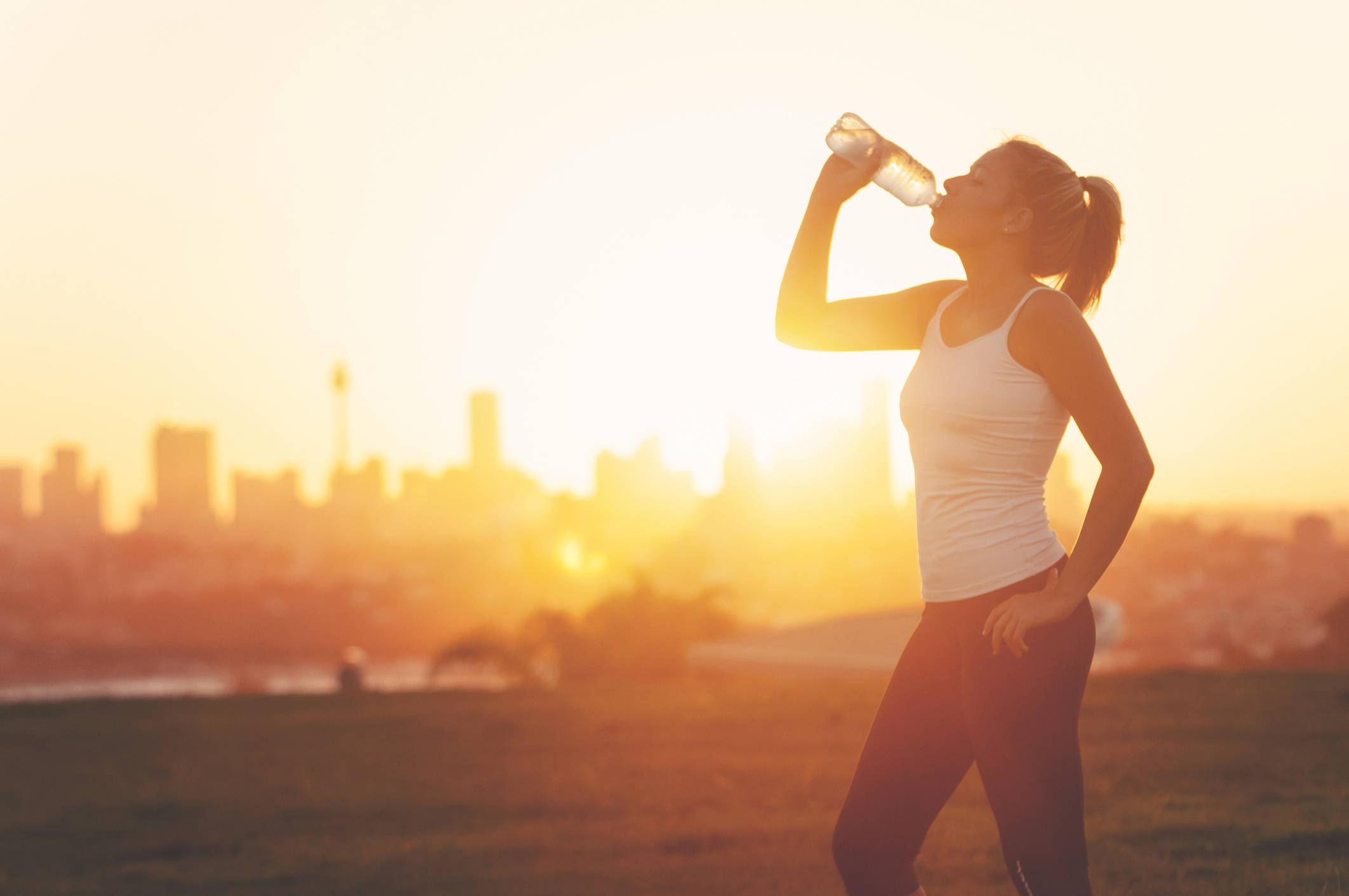 Silhouette of a woman drinking form a cold water bottle. She is exercising at sunset or sunrise. City of Sydney in the background. Copy space.
