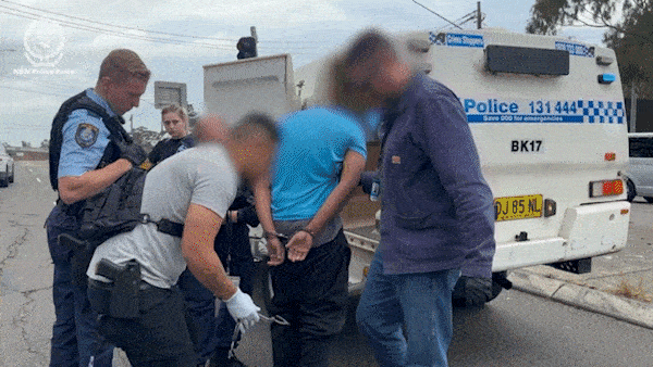A man in a blue shirt with his face blurred being escorted into a police car in handcuffs.