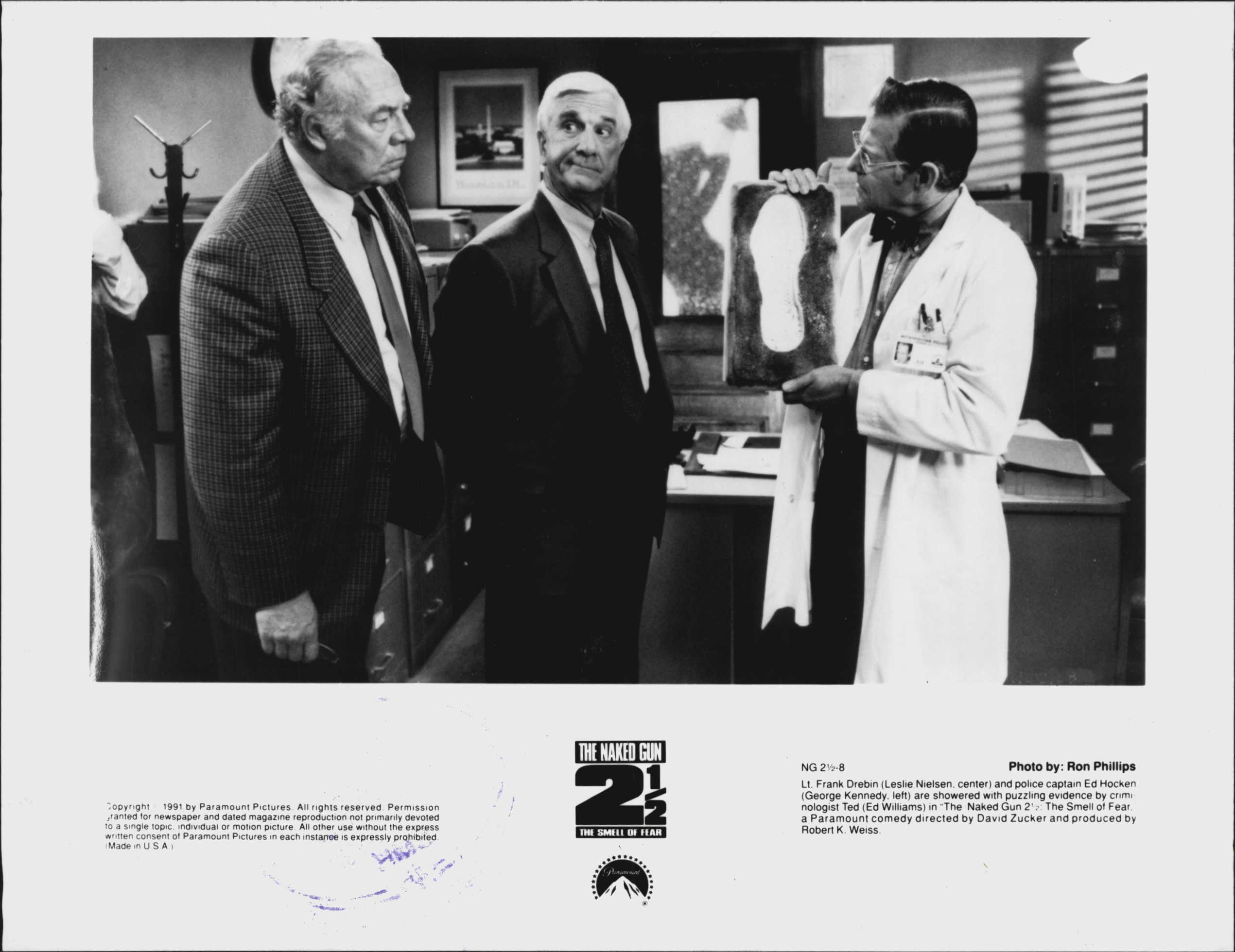 The Naked Gun 2½ The Smile of Fear. Lt. Frank Drebin (Leslie Nielsen. center) and police captain Ed Hocken (George Kennedy, left) are showered with puzzling evidence by Criminologist Ted (Ed Williams) in The Naked Gun 2½The Smell of Fear a Paramount comedy directed by David Zucker and produced by Robert K Weiss. September 12, 1991. (Photo by Ron Phillips, Paramount).