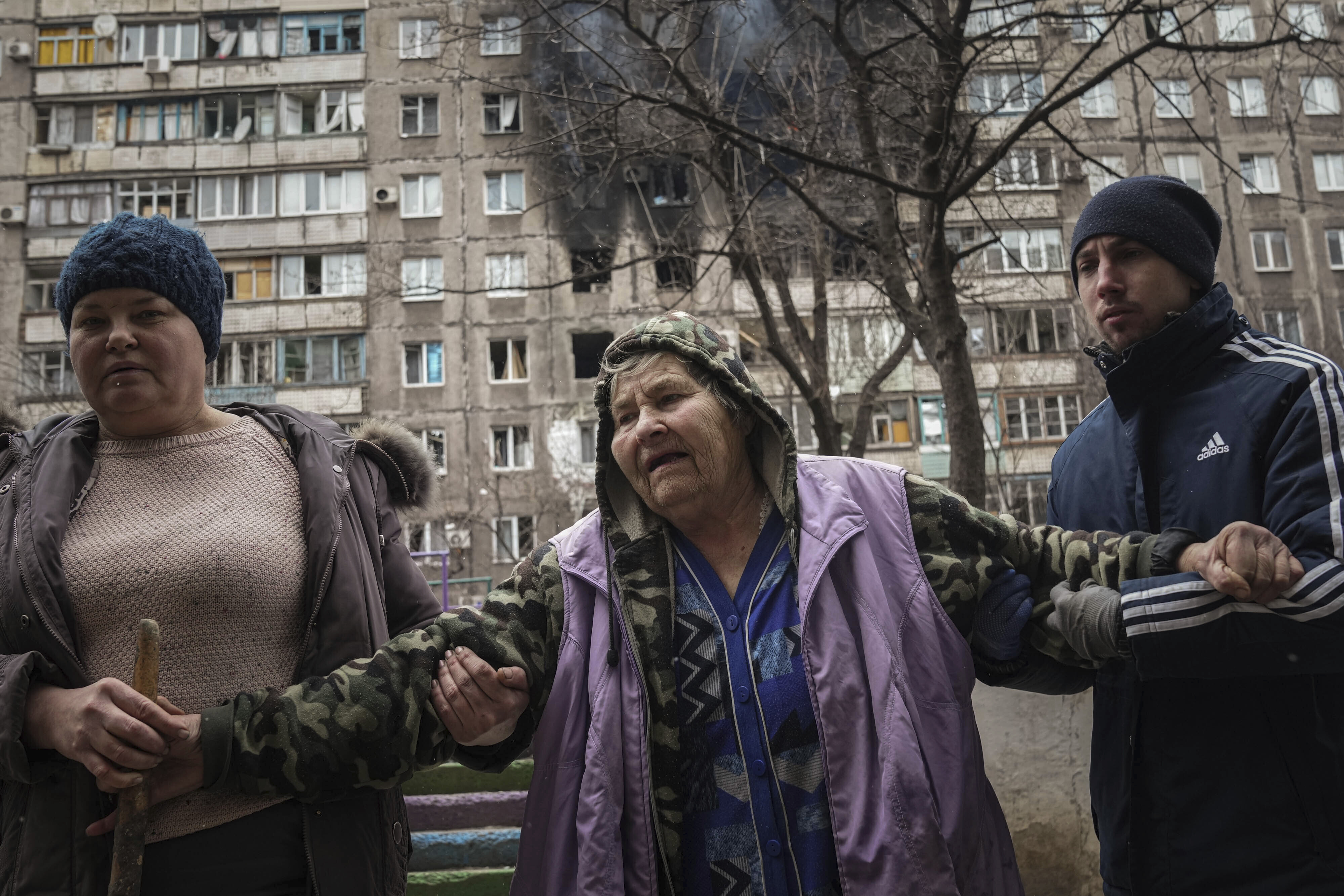 People help an elderly woman to walk in a street with an apartment building hit by shelling in the background in Mariupol, Ukraine, Monday, March 7, 2022.