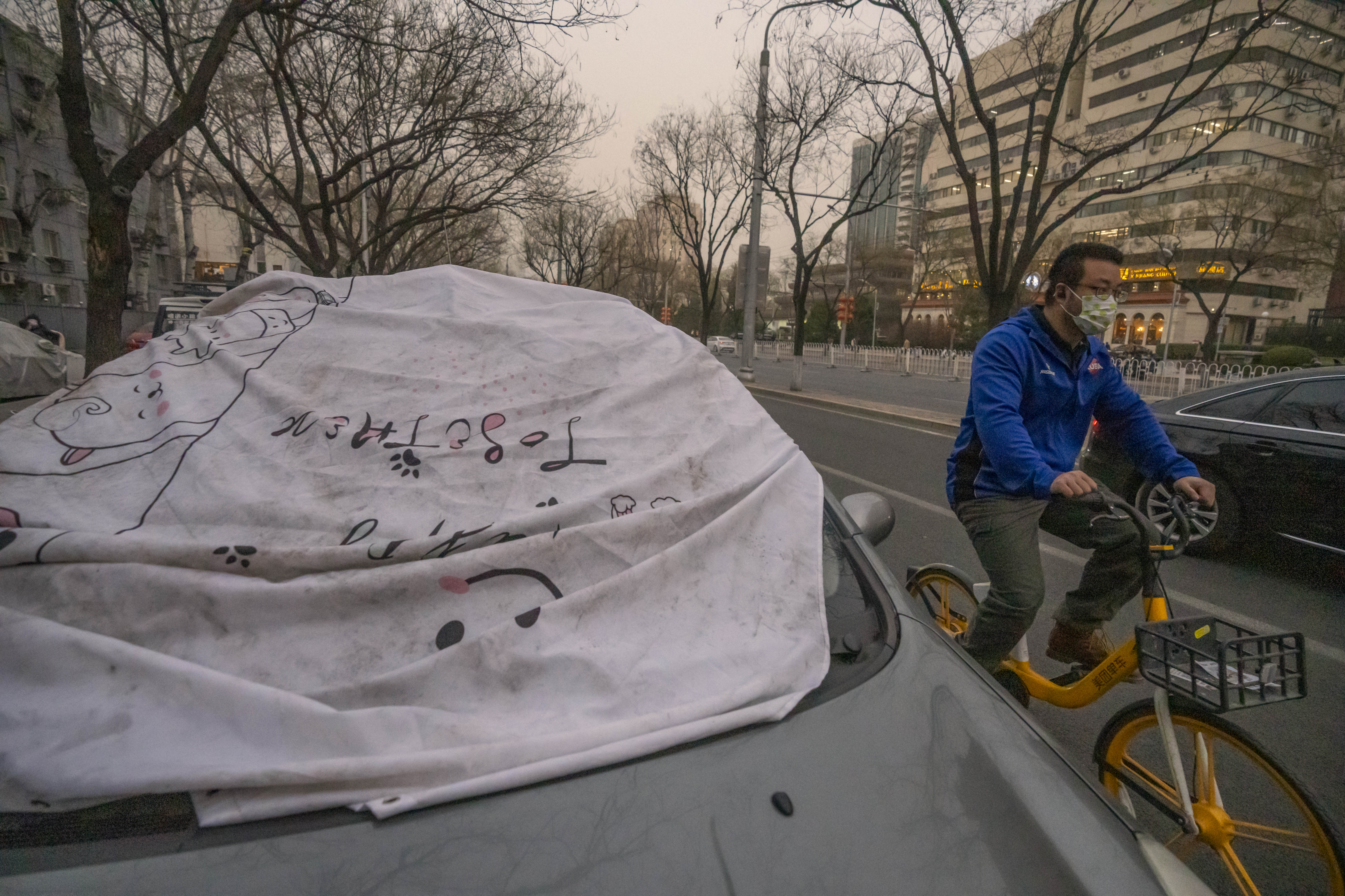 A man wearing a face mask rides past a car with a sheet on the windshield to keep it clean amid dust and haze in Beijing, Friday, March 10, 2023.  