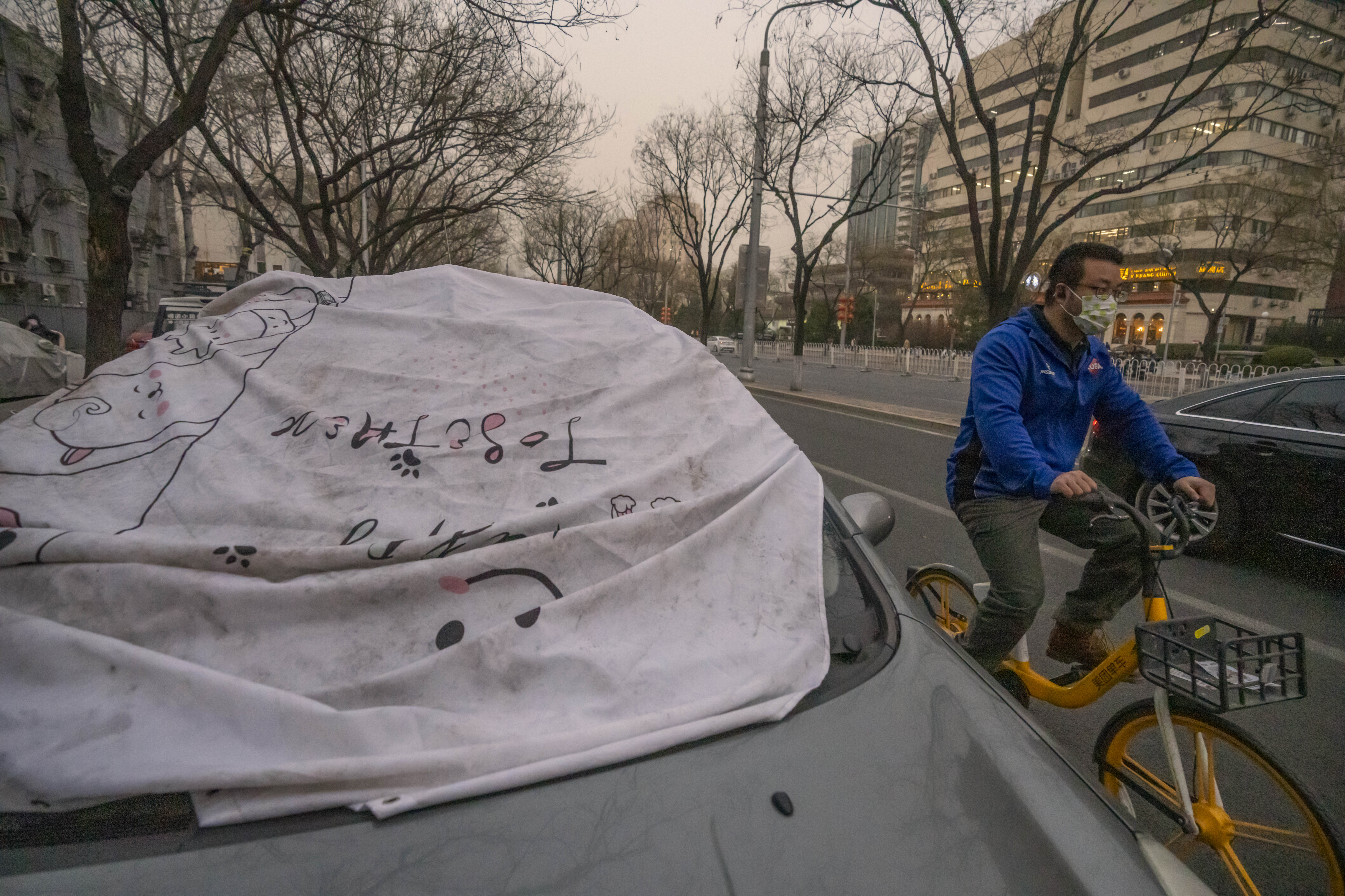 A man wearing a face mask rides past a car with a sheet on the windshield to keep it clean amid dust and haze in Beijing, Friday, March 10, 2023.