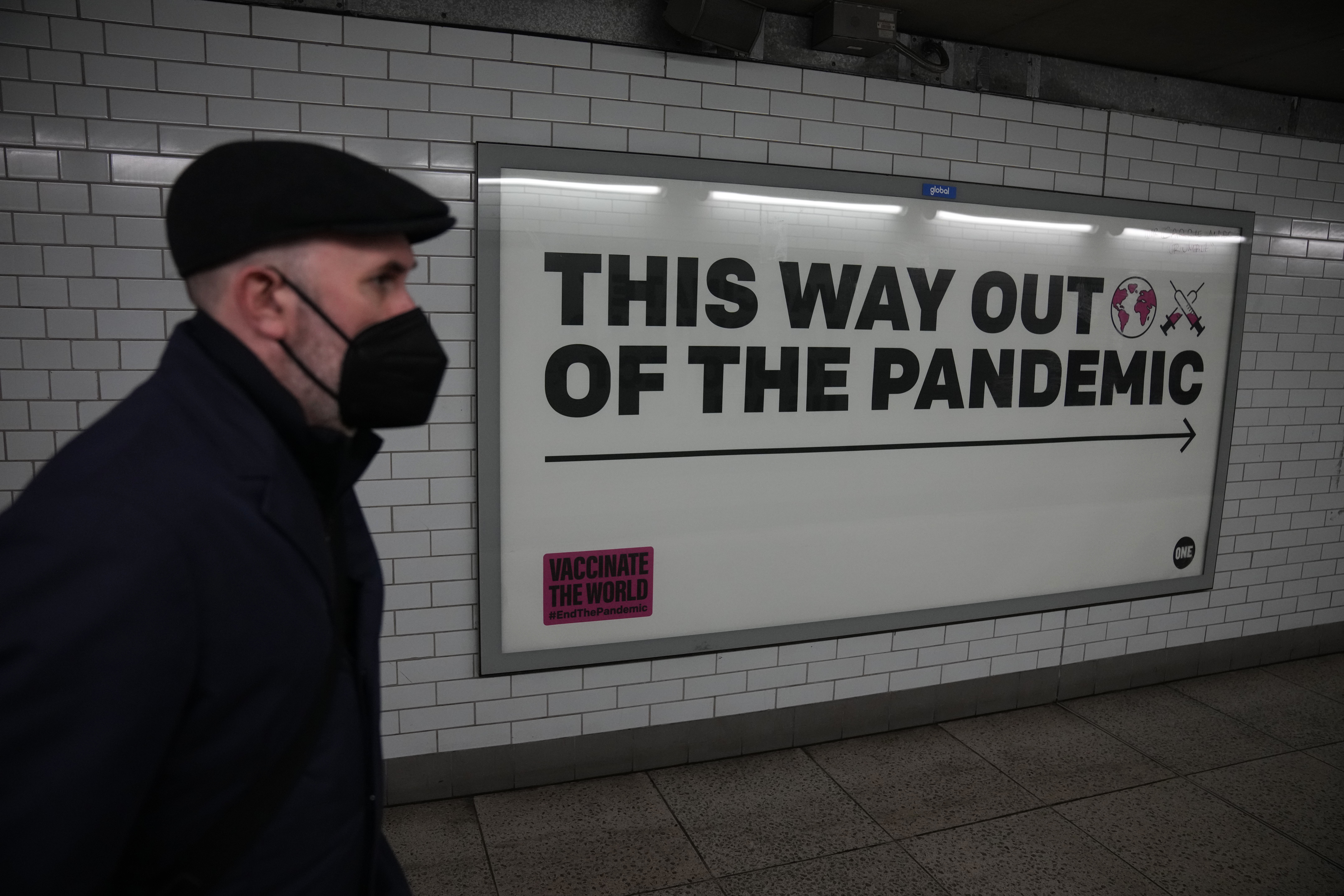 A man wearing a face mask to curb the spread of coronavirus walks past a health campaign poster from the One NGO, in an underpass leading to Westminster underground train station, in London, Thursday, January 27, 2022. 