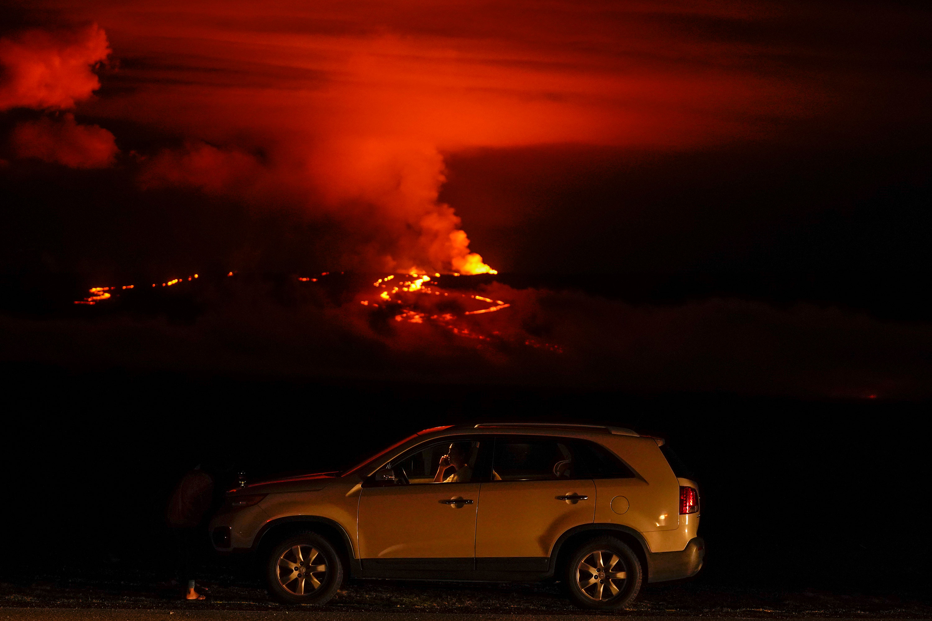 A man talks on a phone in his car alongside Saddle Road as the Mauna Loa volcano erupts Wednesday, Nov. 30, 2022, near Hilo, Hawaii. Hundreds of people in their cars lined Saddle Road, which connects the east and west sides of the island, as lava flowed down the side of Mauna Loa and could be seen fountaining into the air on Wednesday. 