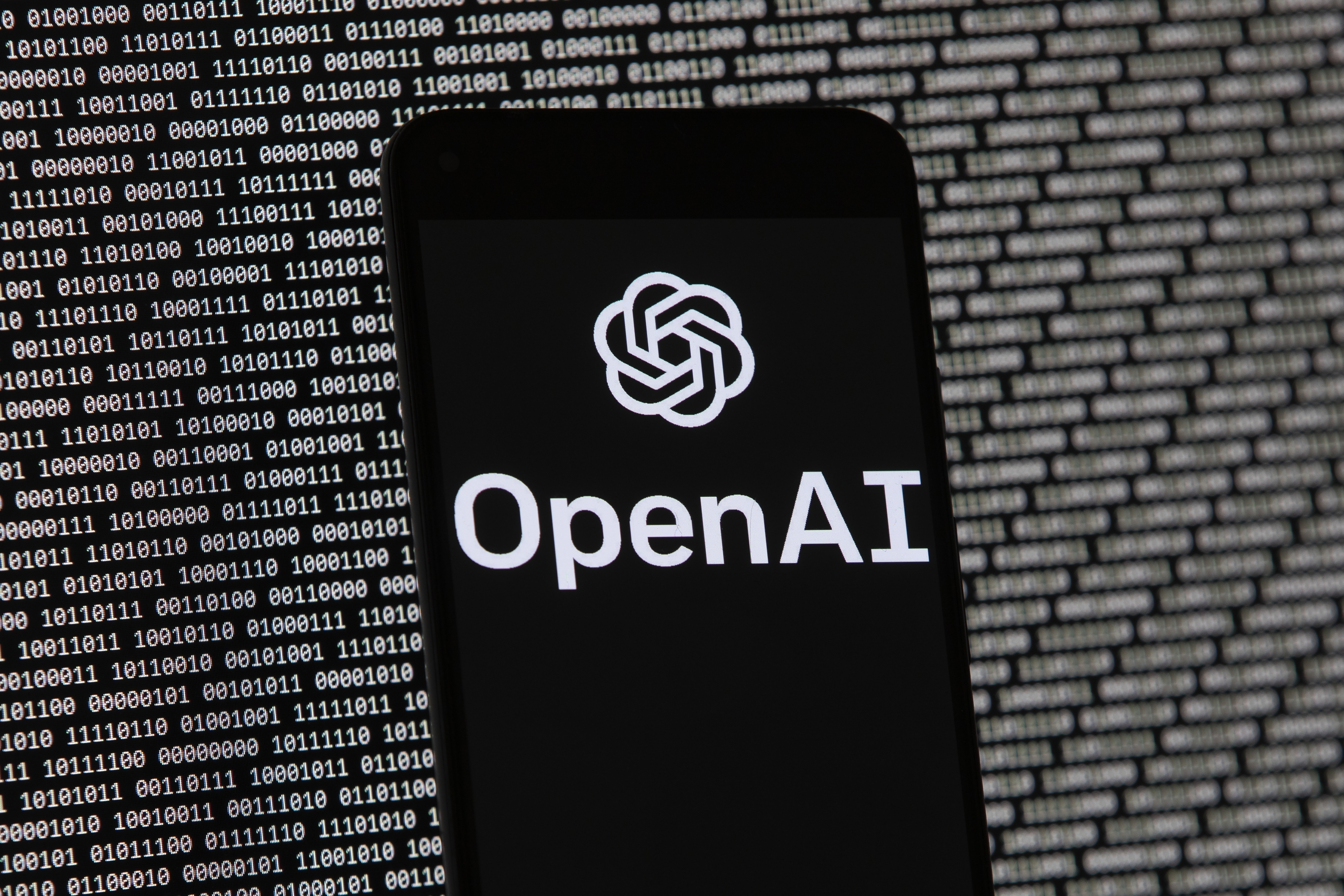 The OpenAI logo appears on a mobile phone in front of a computer screen with random binary data, March 9, 2023, in Boston. (AP Photo/Michael Dwyer, File)