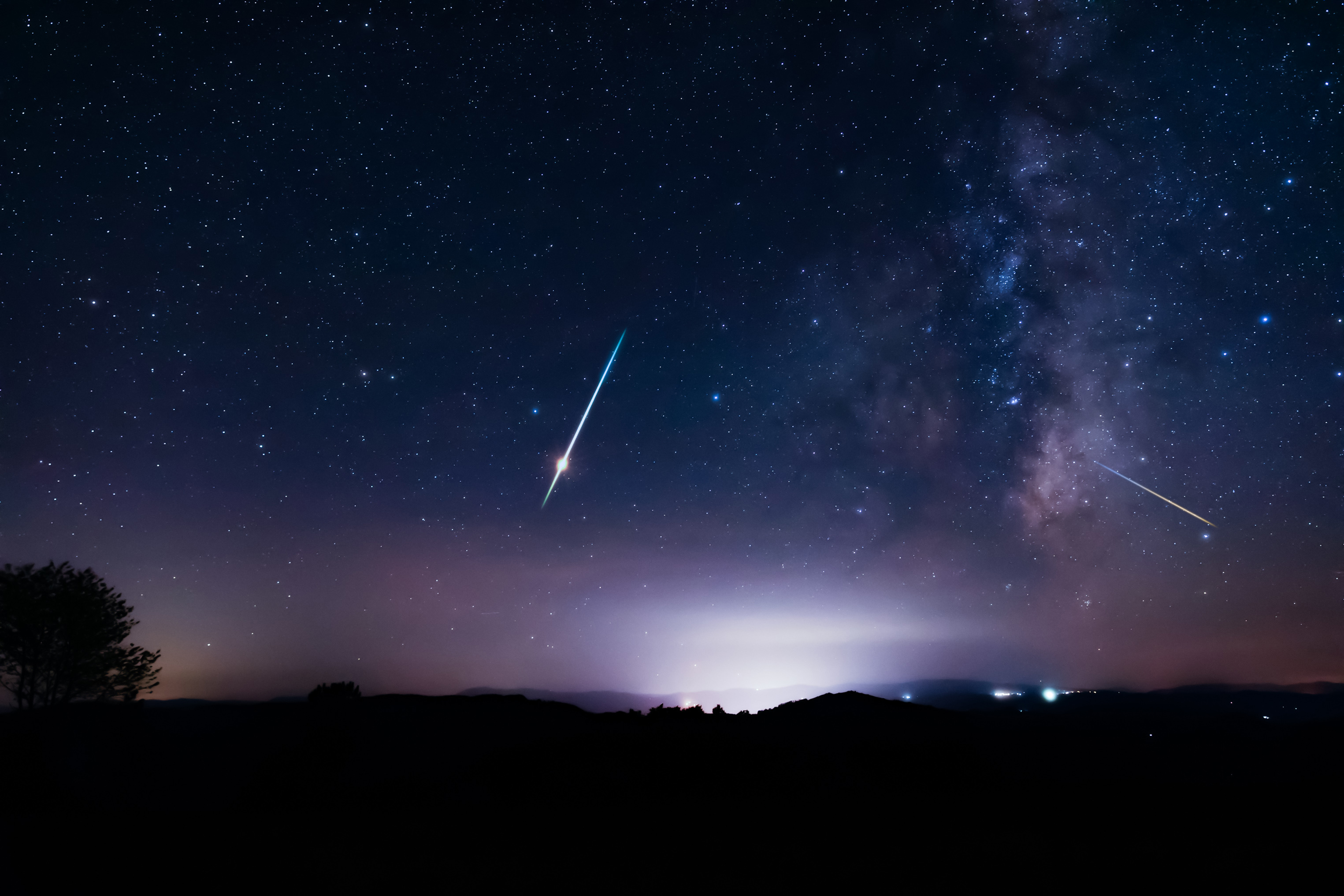 Milky Way stars with meteor shower trails and countryside silhouettes.