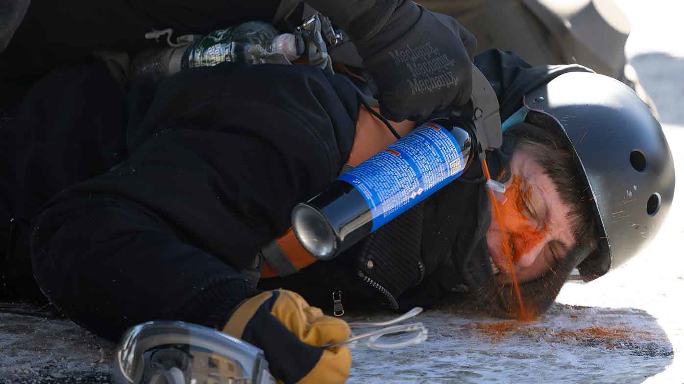 An ICE agent sprays pepper spray directly into the eyes of a protester who is pinned to the ground.