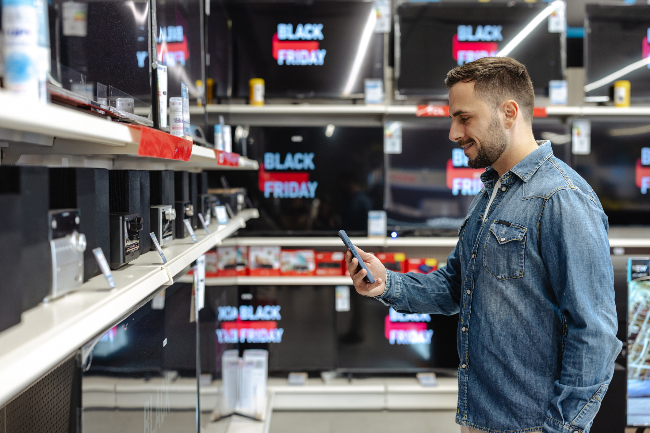 A young man is browsing electronics in a store during a Black Friday sale, holding his smartphone.