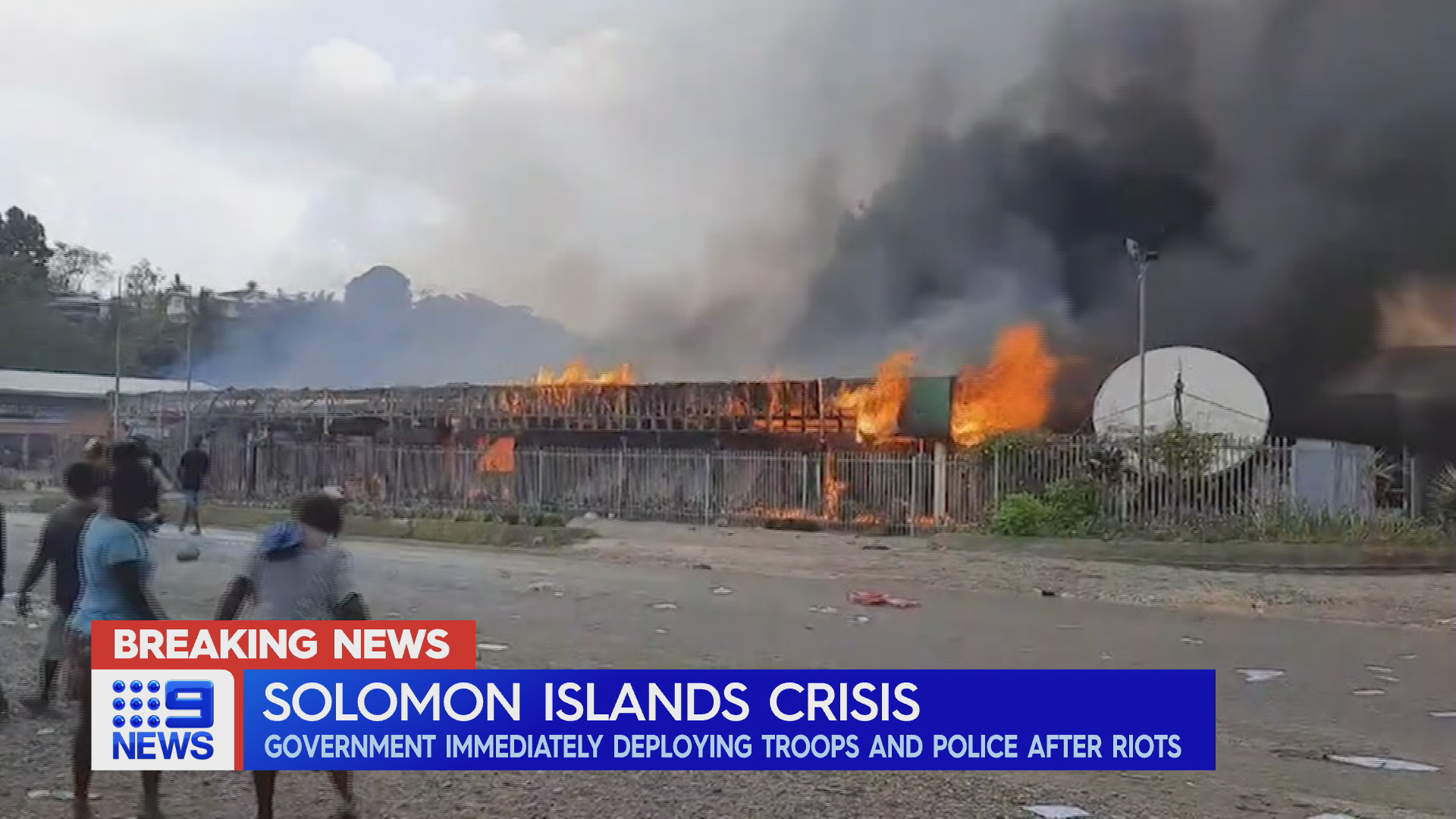 Wild flames tear through a property as people, potentially demonstrators, linger nearby.