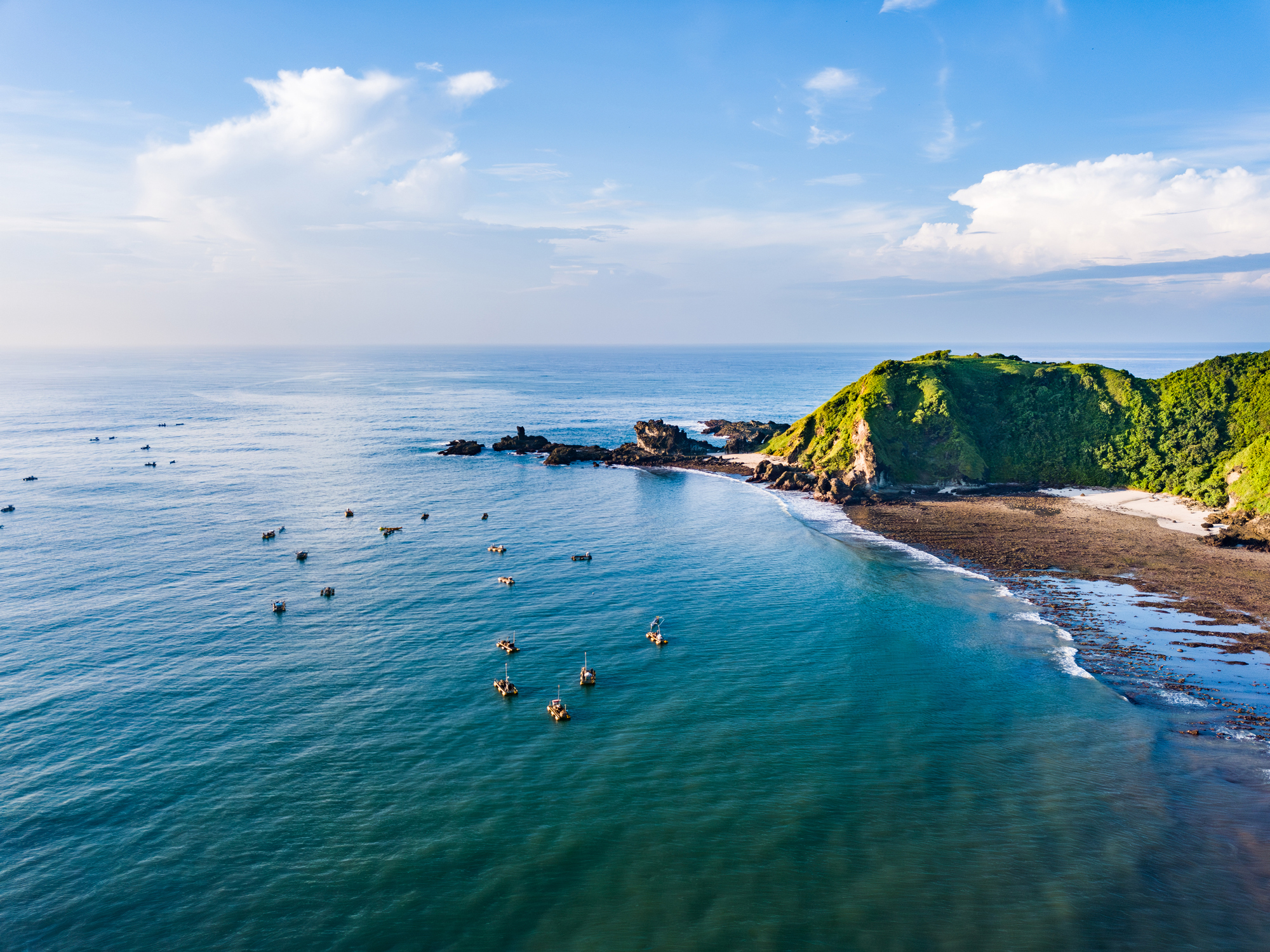 A lush green hillside meets the clear blue waters of the ocean. The rocky shoreline is visible, adorned with various shapes and sizes of rocks. In the distance, a cloudy sky paints a serene backdrop. Shot taken on Lombok.
