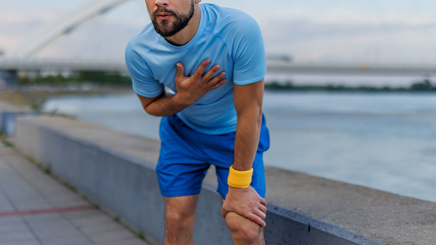 A man in a light blue shirt and blue shorts is exercising outdoors by the river, holding his chest and bending over, with a bridge in the background, conveying fatigue.