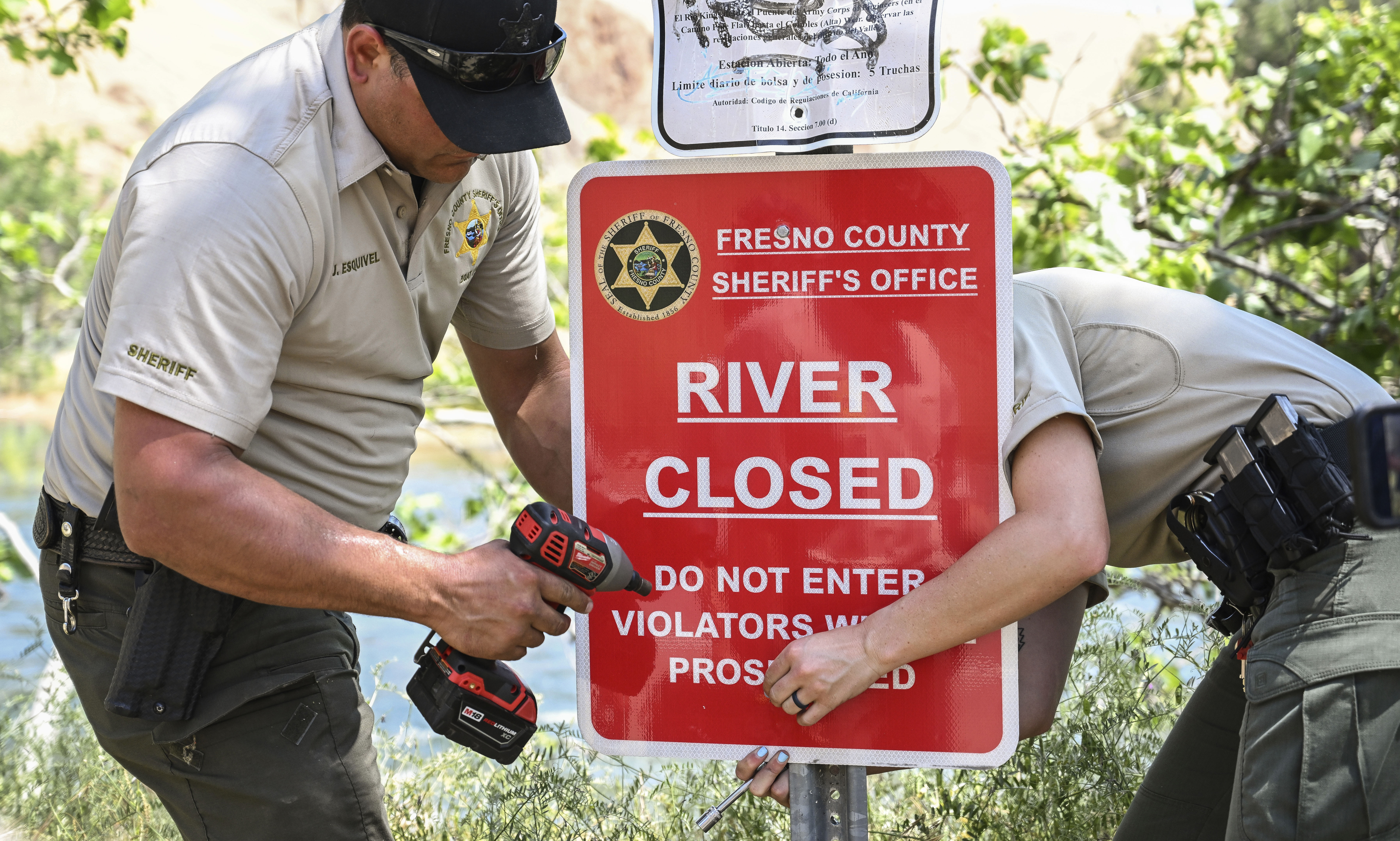 Fresno County Sheriff's Office reservists attach a sign to a post to alert people that the the Kings River is closed due to dangerously high water levels, on Monday, May 22, 2023, near Sanger, Calif. The body of a 4-year-old boy was recovered Monday from the surging California river that is closed to recreational use, a day after his 8-year-old sister died when the siblings were swept away by the current, authorities said. (Craig Kohlruss/The Fresno Bee via AP)