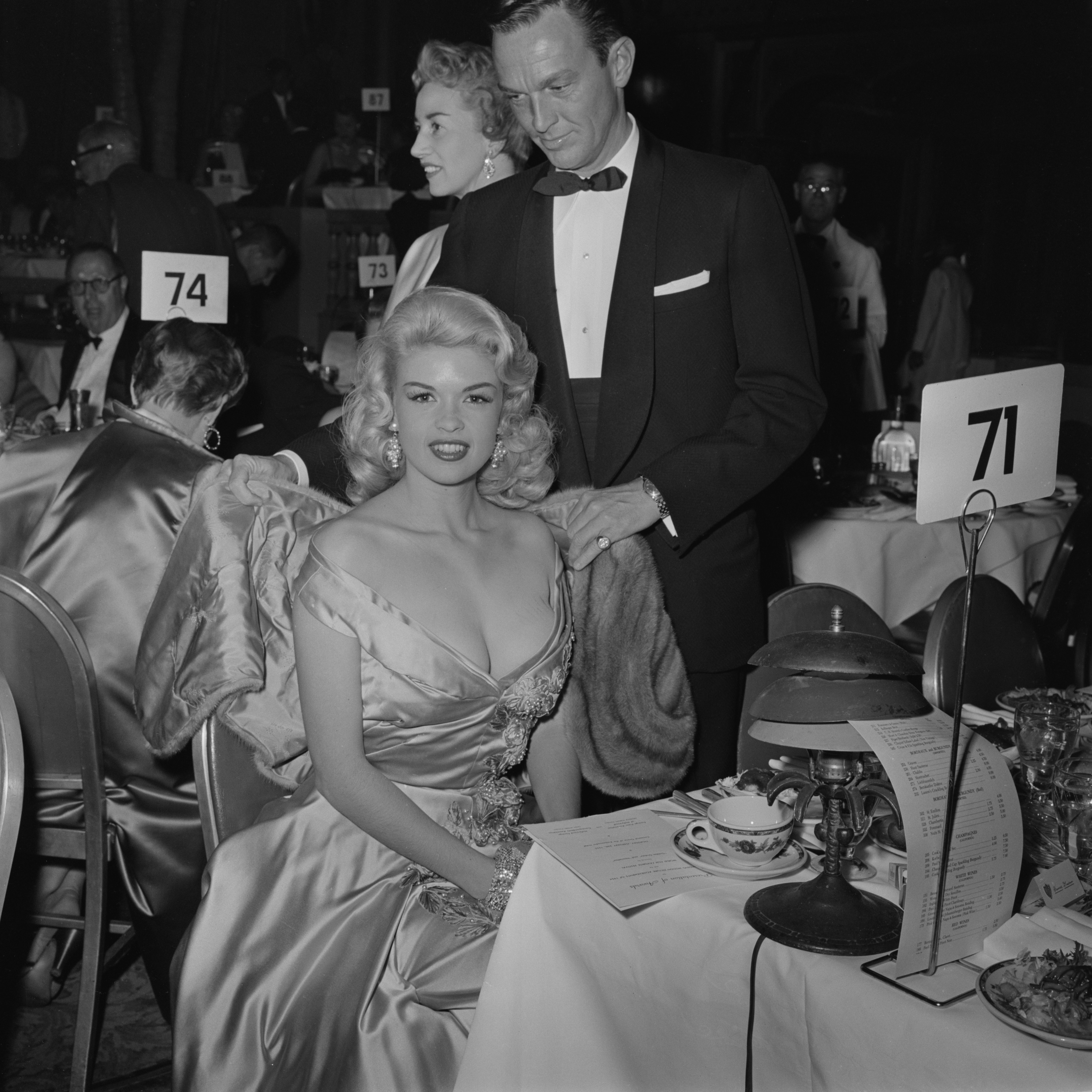Jayne Mansfield sitting at a table as a man in a tuxedo puts a fur wrap around her shoulders, at the 12th Golden Globe Awards at the Ambassador Hotel in Los Angeles, California, 24 February 1955.
