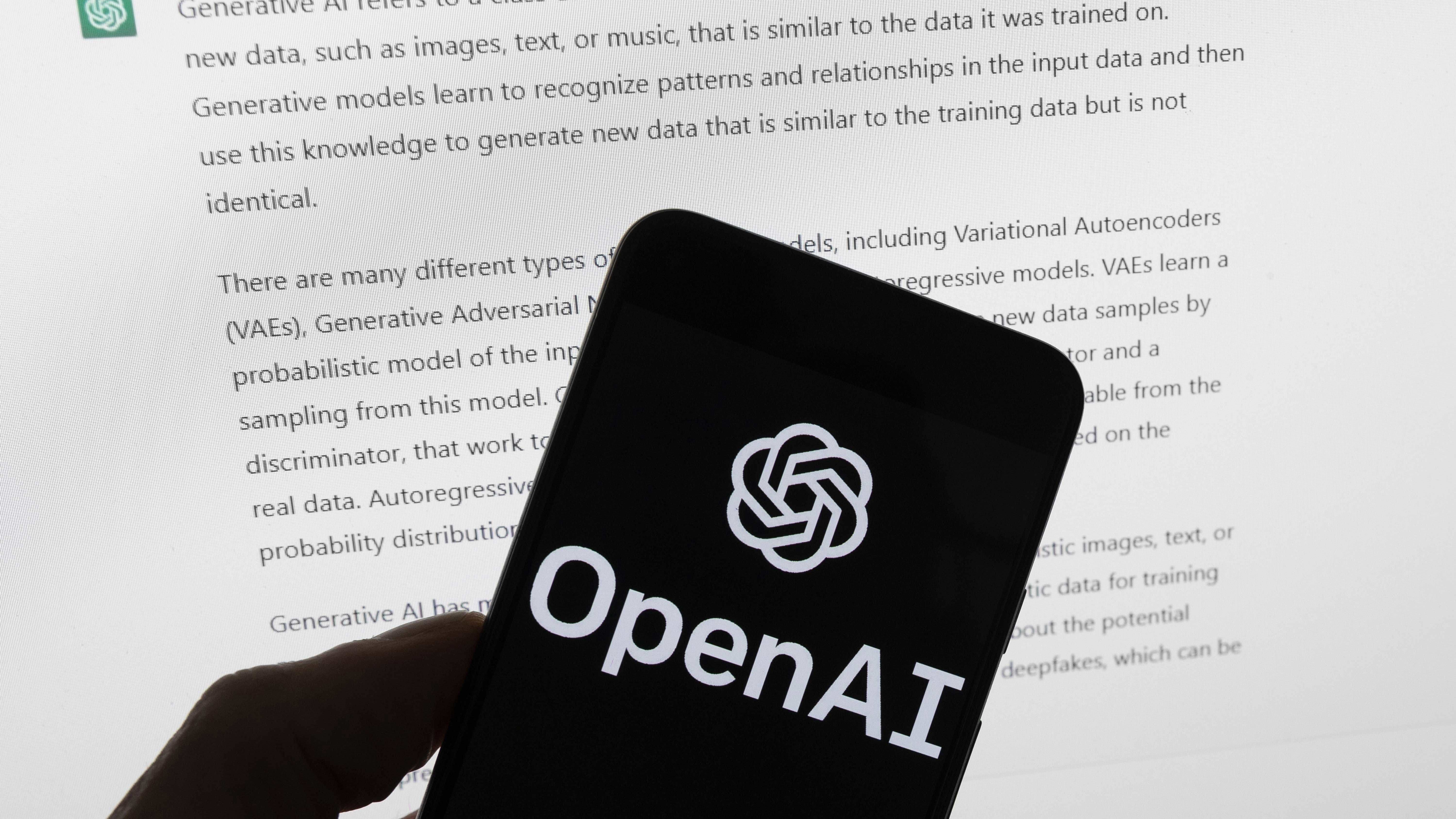 El logotipo de OpenAI se ve en un teléfono móvil frente a una pantalla de computadora que muestra la salida de ChatGPT.