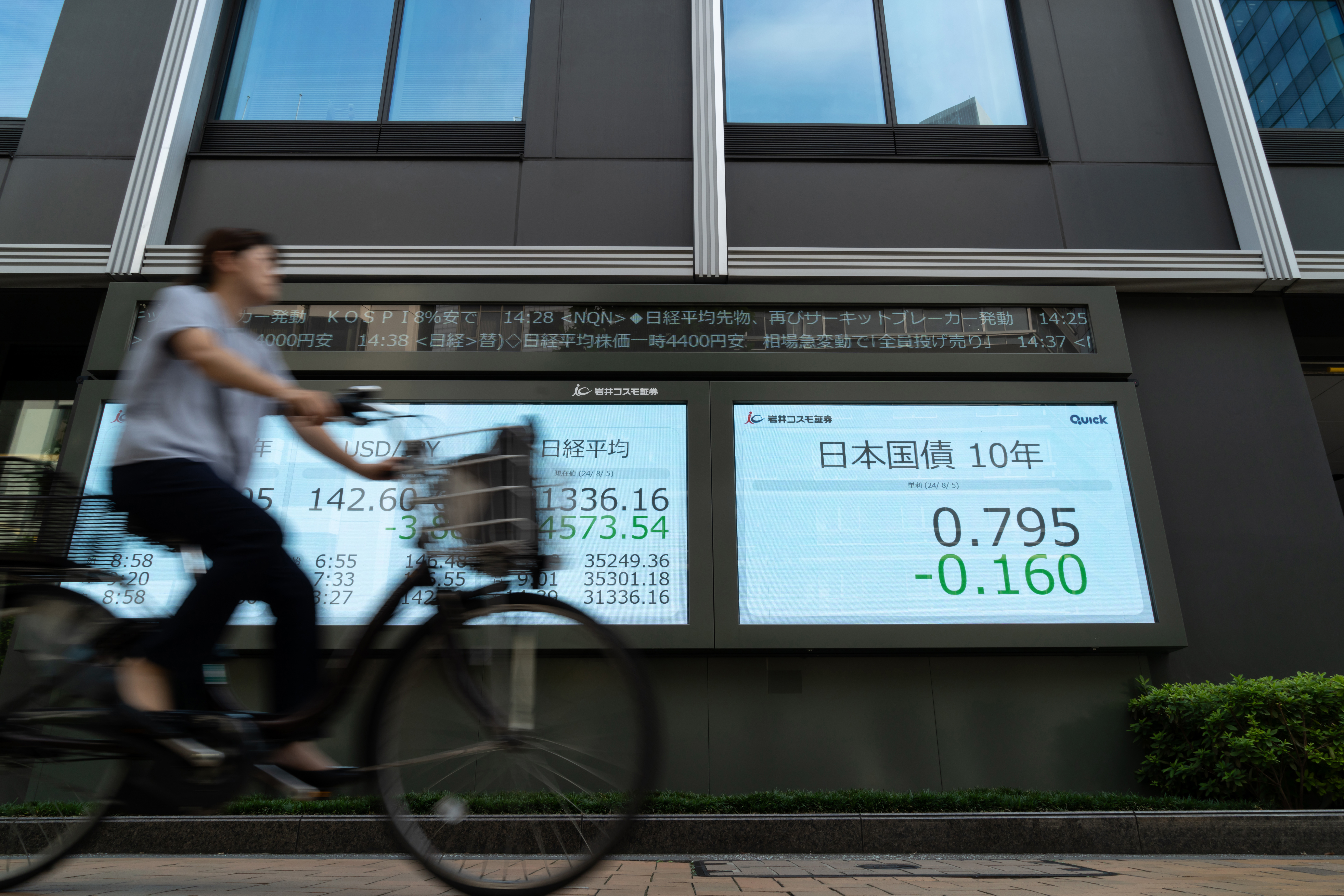 A pedestrian cycles past monitors displaying the Nikkei 225 Stock Average figure and other financial data outside a securities firm on August 5 in Tokyo, Japan.