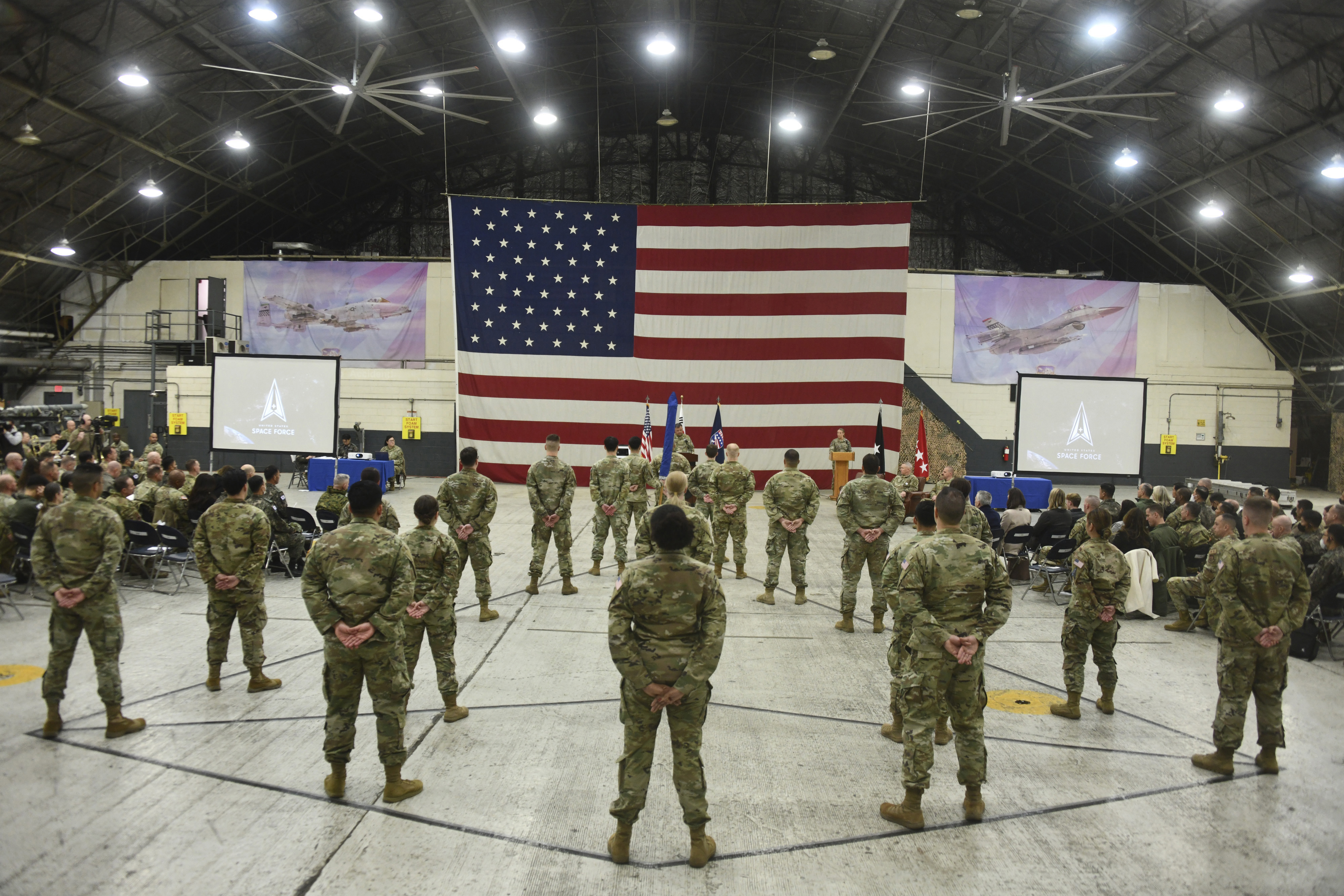 Soldados estadounidenses de pie en una sala en la ceremonia de activación de las Fuerzas Espaciales de los Estados Unidos con una bandera estadounidense frente a ellos