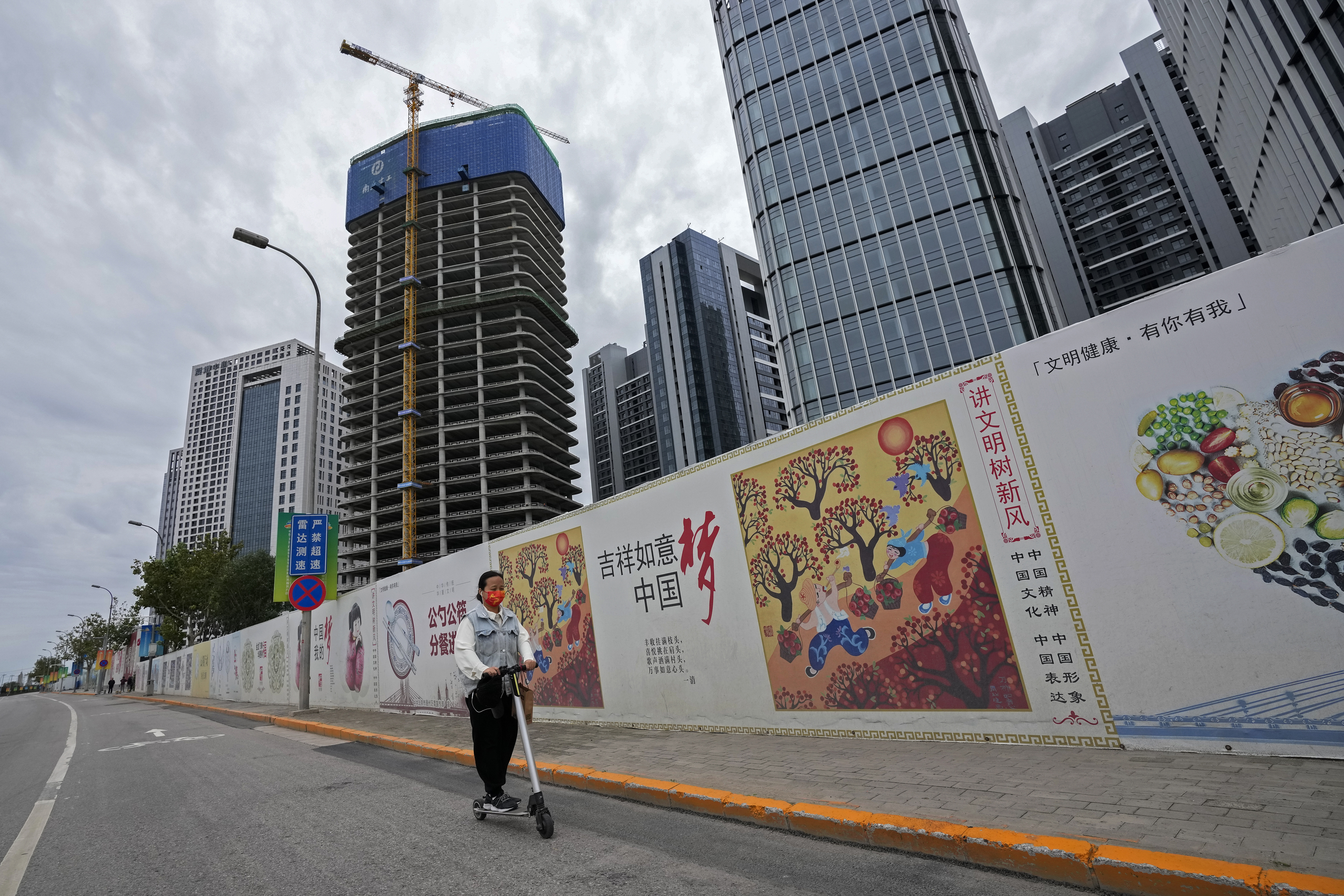 A girl wearing a face mask to help protect from the coronavirus ride a scooter past by Chinese government's propaganda "China Dream" billboard on display along a commercial office buildings under construction in Tongzhou, outskirt of Beijing.