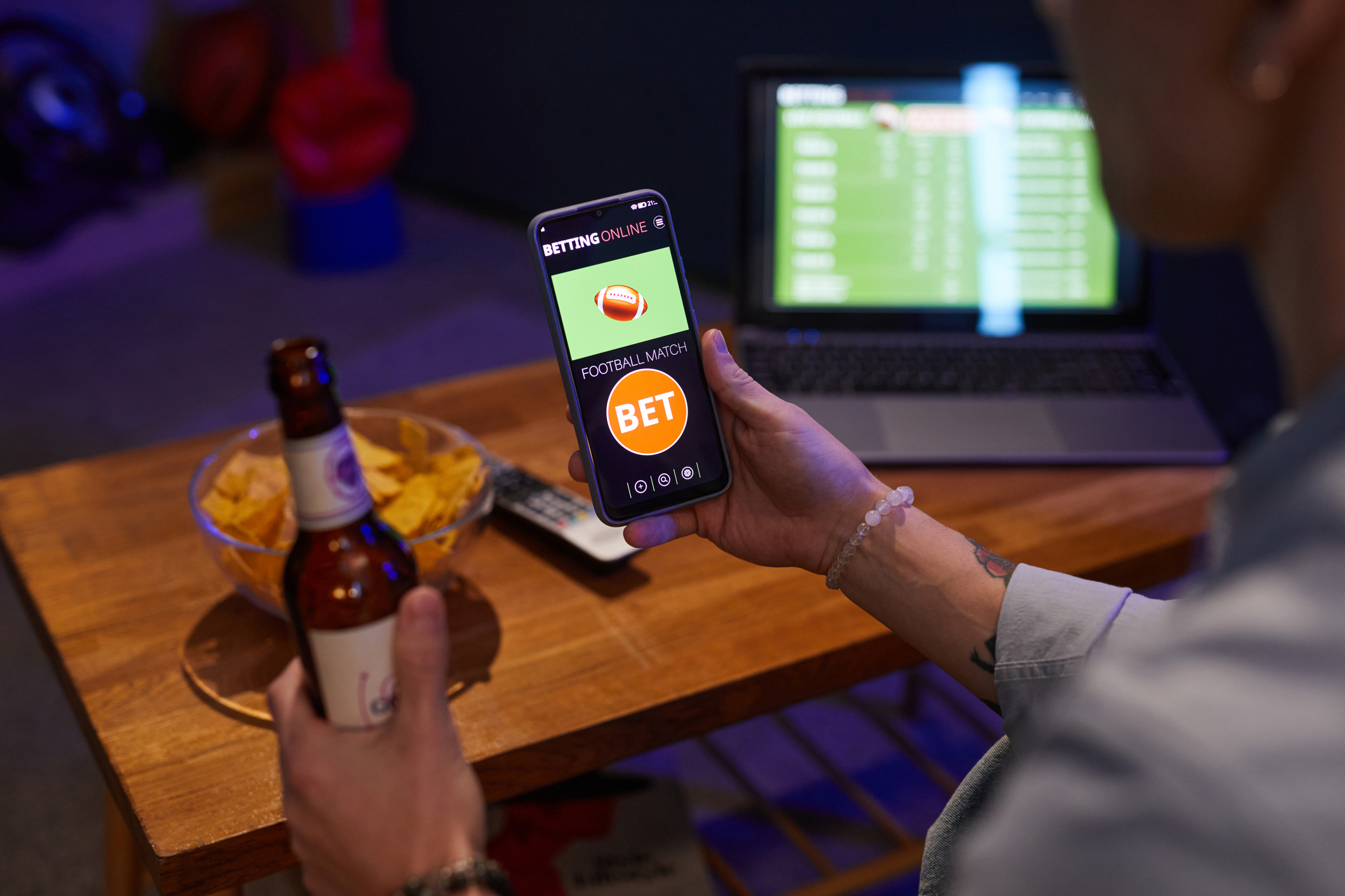 Person holding smartphone showing betting online app while drinking beer in living room, with laptop and snacks on wooden table