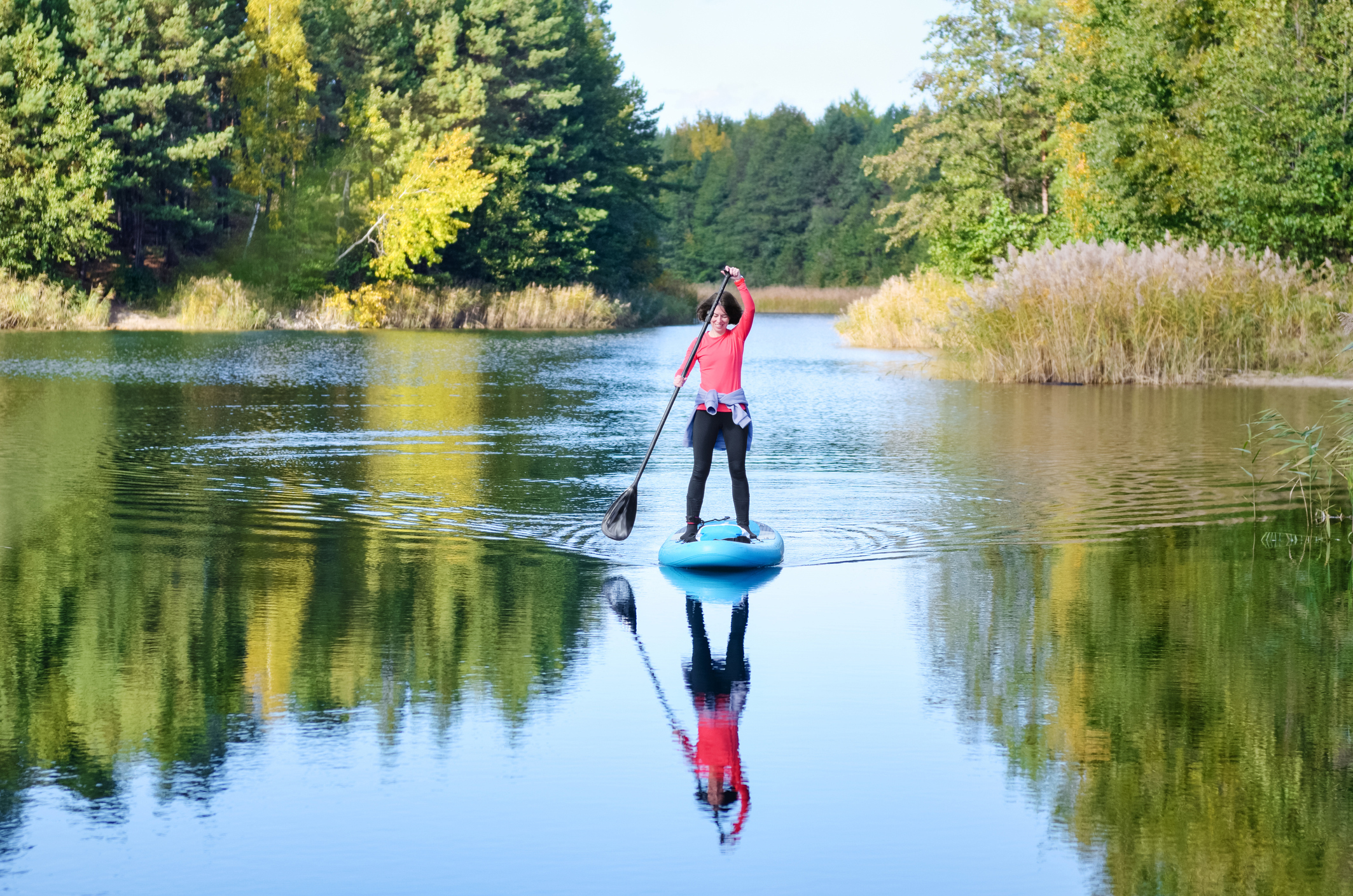 Active woman paddling SUP board on beautiful lake, autumn forest landscape and nature on background, stand up paddling water adventure outdoors, sport and healthy lifestyle concept