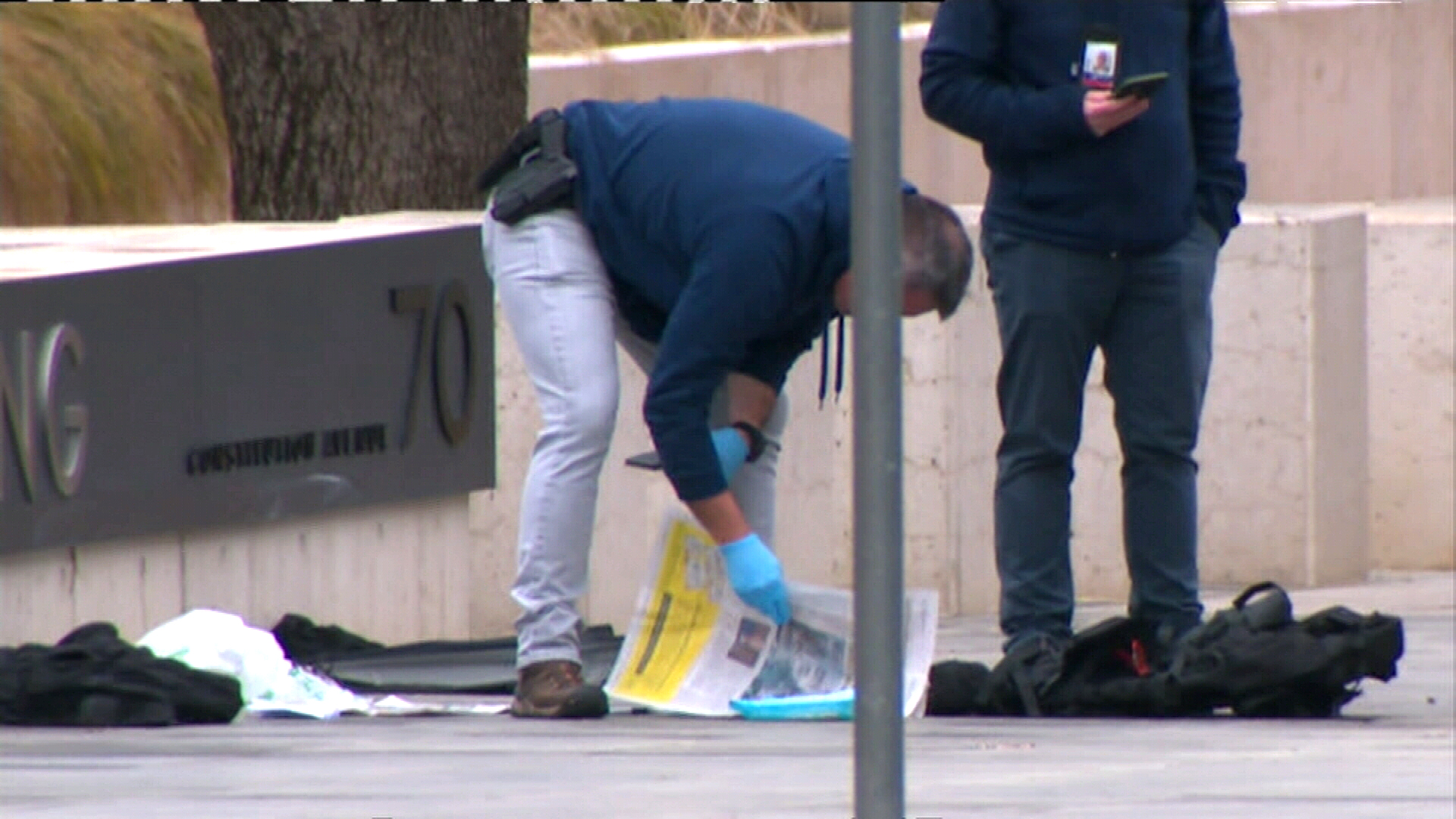An armed, plain clothes officer looks over items on the ground, in front of the ASIO headquarters.