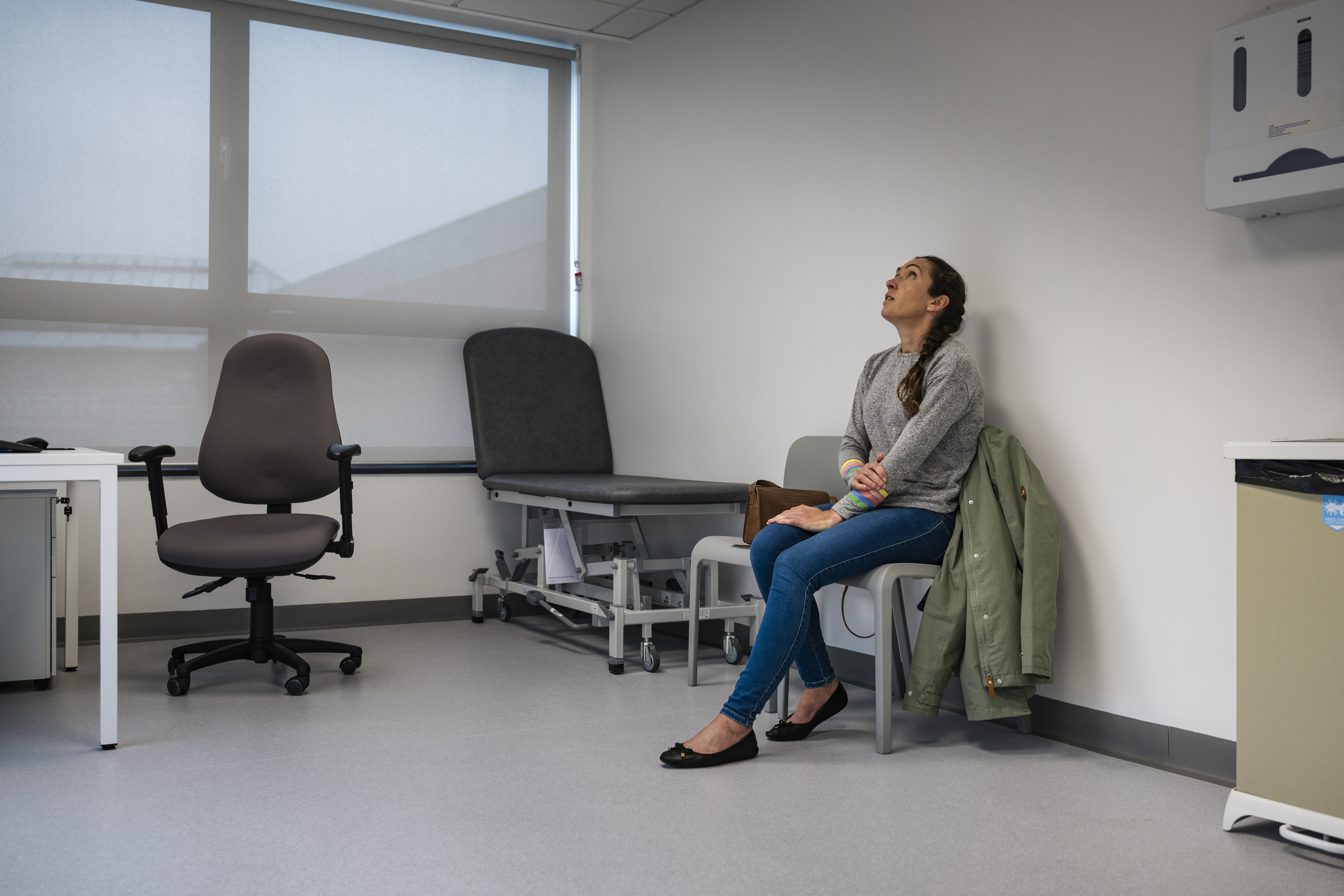 Full length wide view of a mid adult woman sitting in a doctors examination room waiting to be seen, she is holding her arm and looking up, feeling worried. She is wearing casual clothing at the hospital in Newcastle, England.