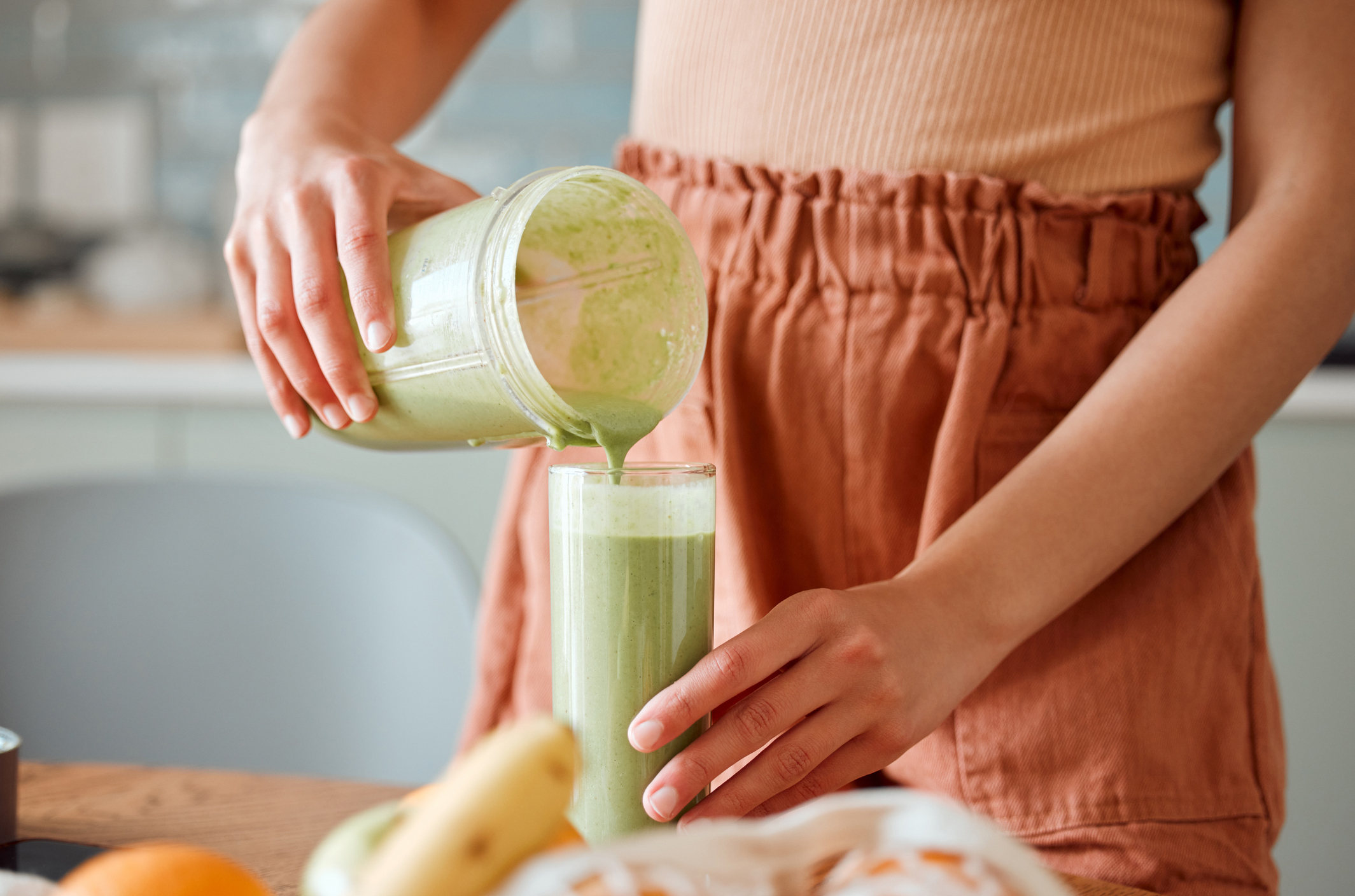 Woman pouring healthy smoothie in a glass from a blender jar on a counter for detox. Female making fresh green fruit juice in her kitchen with vegetables and consumables for a fit lifestyle. stock image