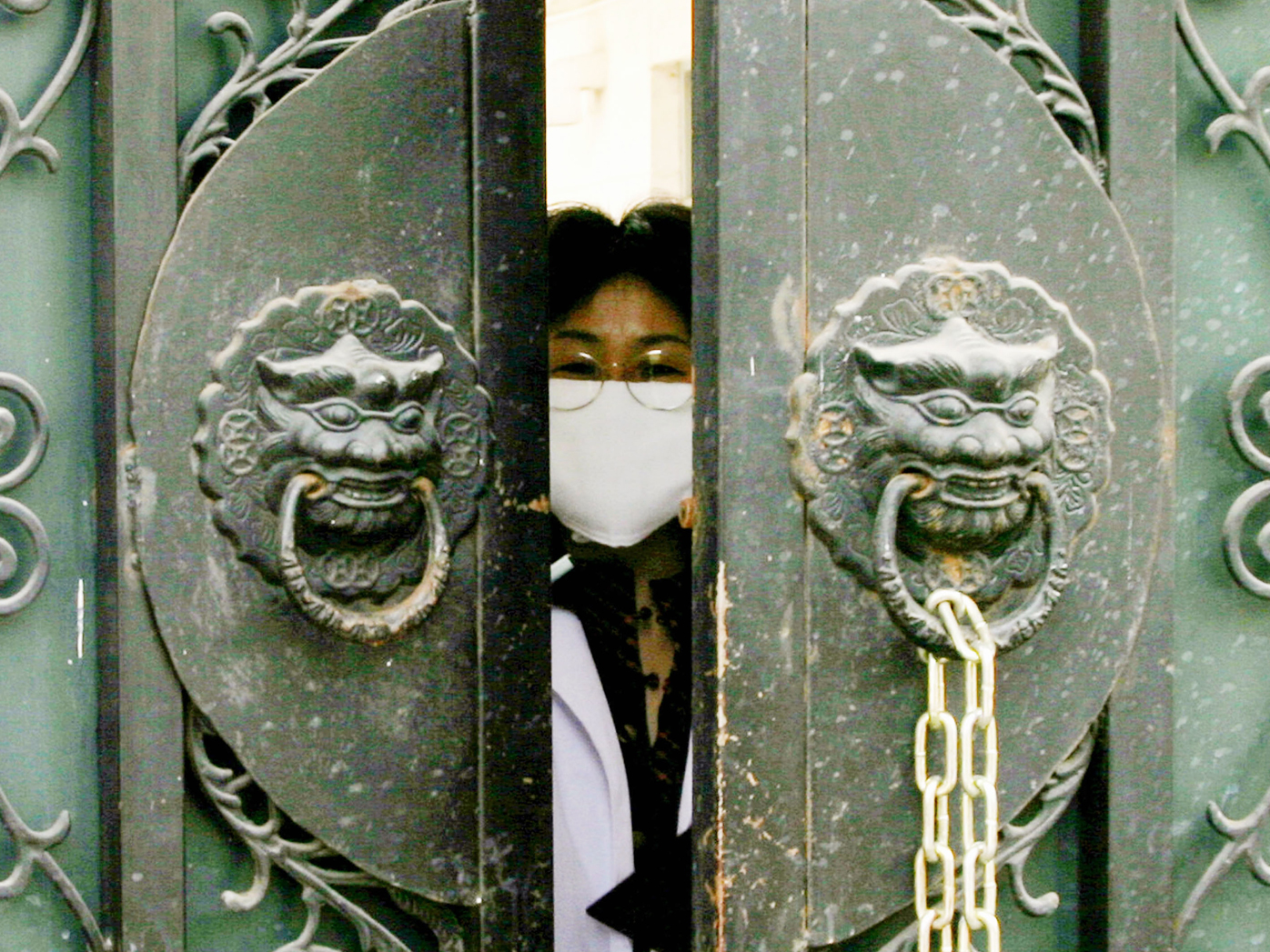 In this 2004 photo, a quarantined medical worker, wearing a face mask to protect against the Severe Acute Respiratory Syndrome (SARS) virus, peeks out of the entrance gate of the national institute of virology to pick up clothes and lunch boxes in Beijing, China.